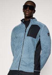 Jachetă fleece de culoare albastru deschis cu țesătură texturată, buzunar cu fermoar negru și panou lateral negru. Închidere cu fermoar și design cu guler înalt.
