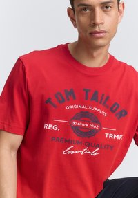 Rød bomulds T-shirt med korte ærmer, der har et grafisk print i marineblå og hvid tekst, som viser "TOM TAILOR" og "PREMIUM KVALITET."