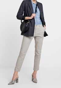 Pantalon beige fuselé, chemise en denim clair, blazer à carreaux bleu marine et chaussures à talons blocs argentées à motif serpent. Sac rond noir en accent.