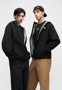 Δύο μοντέλα φορούν μαύρα μπουφάν με φερμουάρ και το λογότυπο The North Face· το ένα φοράει μαύρο παντελόνι, το άλλο καφέ παντελόνι και καπέλο με την πίσω πλευρά μπροστά.