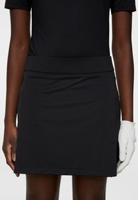 Svart idrotts kjol med bred midjeband och slät tyg. Har en ren design, knä-lång klippning, och är parat med en vit golfhandske.