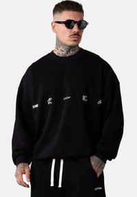 Sweat-shirt noir à col rond et épaules tombantes, avec des logos blancs. Le matériau semble doux, avec une coupe décontractée et des poignets côtelés.