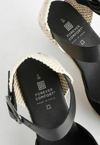 Sandali in pelle nera con cinturini intrecciati color crema, suole testurizzate con il marchio "Forever Comfort", prodotti in Spagna. Taglia 39.