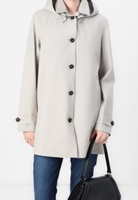 Gabardina gris claro con capucha, que presenta seis botones negros, puños ajustables y una textura suave; combinada con un bolso negro.