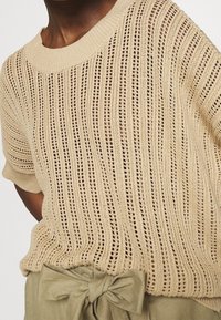 Väga beige kootud topp, millel on lõtv istuvus, vertikaalne avatud kudumismuster, ümar kaelus ja lühikesed varrukad.