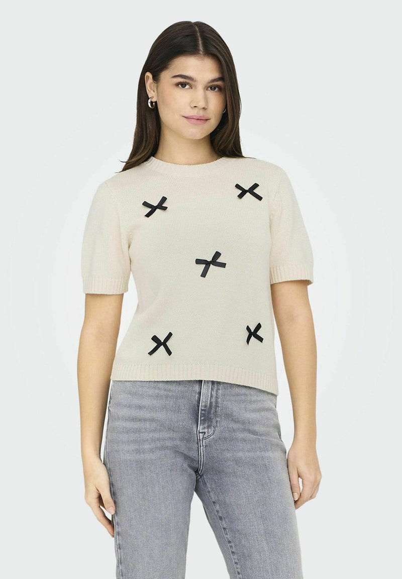 Lichtbeige korte mouw trui met een geribbelde zoom, voorzien van zwarte linten kruis applicaties die gelijkmatig over de voorkant zijn verdeeld.