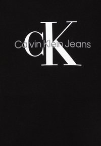 Tissu noir avec un logo "CK" blanc bien en vue et le texte "Calvin Klein Jeans" en gris. Design simple avec un bon contraste de texte.