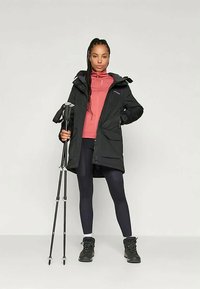 Chaqueta impermeable negra con capucha, capa intermedia rosa, leggins negros y botas negras. El usuario sostiene un bastón de senderismo y está de pie sobre un fondo claro.