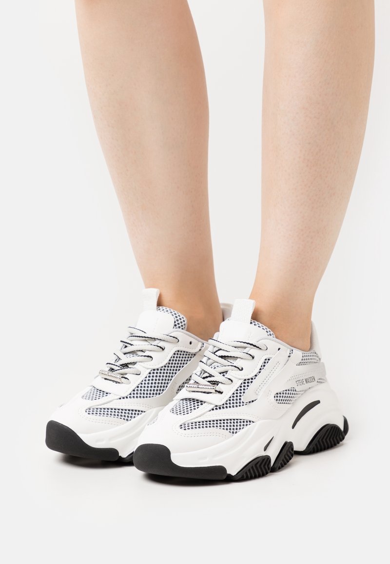 Baskets épaisses blanches et noires avec des détails en mesh portées sur des jambes nues sur un fond blanc uni.
