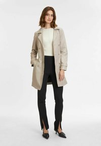 Trench coat beige chiaro con bottoni doppi, revers, tasche laterali e una vestibilità rilassata. Indossato sopra un top bianco a coste e pantaloni neri con fessure.