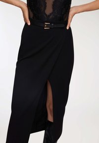Donna che indossa una gonna nera a vita alta con spacco frontale, cintura nera con fibbia dorata, top in pizzo nero e stivali di pelle neri.