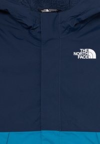 Námořná a tyrkysová outdoorová bunda s kapucňou, so bielym logom The North Face na hrudi.
