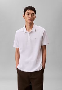 Muž stojící s rukama v kapsách, oblečený v bílé krátkém rukávu polo tričku a tmavě hnědých kalhotách na jednobarevném šedém pozadí.