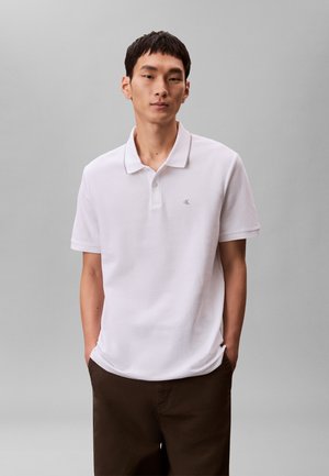 Bărbat în picioare cu mâinile în buzunare, purtând un tricou polo alb cu mânecă scurtă și pantaloni maro închis, pe un fundal gri simplu.