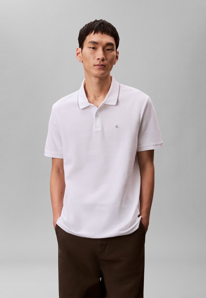 Muž stojící s rukama v kapsách, oblečený v bílé krátkém rukávu polo tričku a tmavě hnědých kalhotách na jednobarevném šedém pozadí.