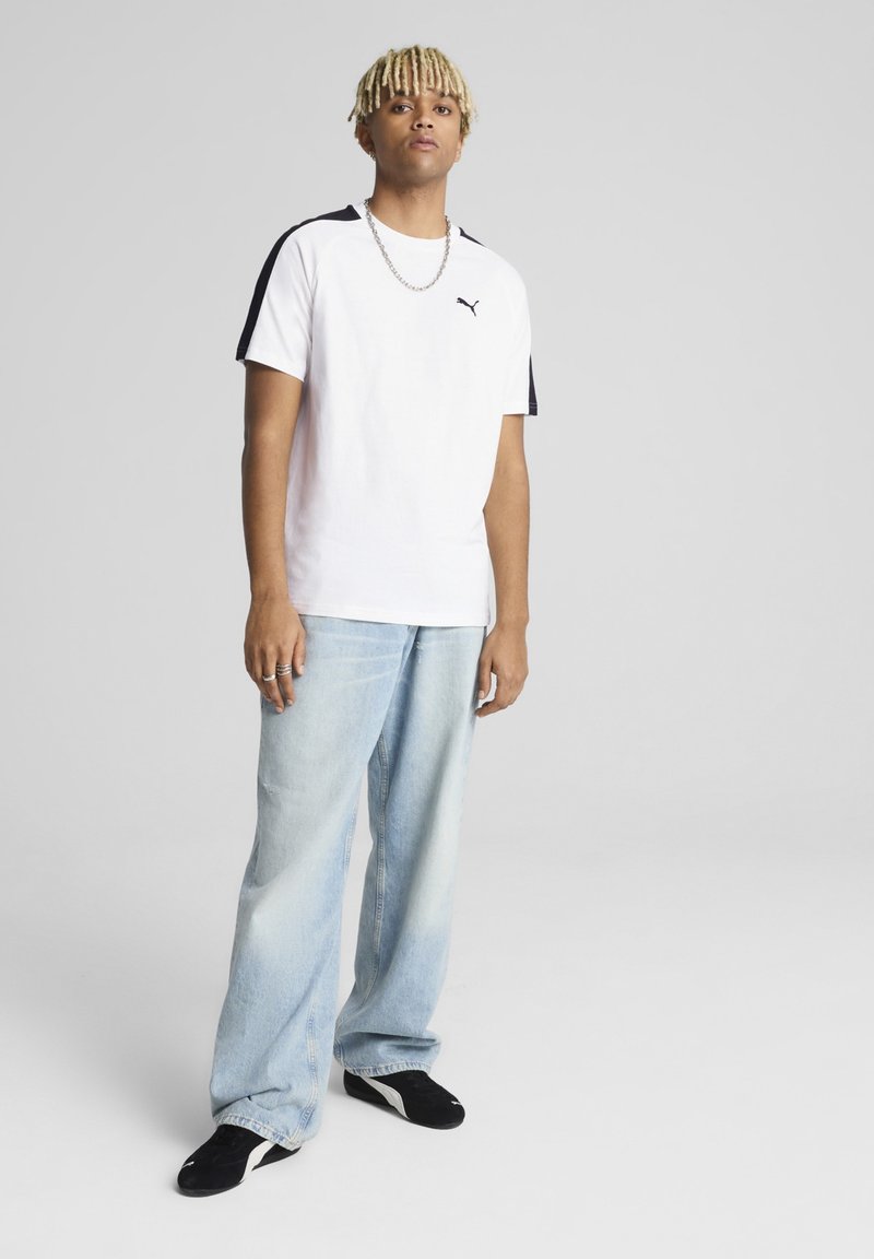 Camiseta blanca de manga corta con acentos laterales negros y el logo de Puma, combinada con jeans de corte holgado azul claros y zapatos negros.