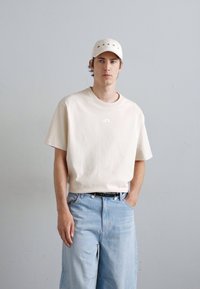 Cream short-sleeve t-shirt with a small logo on the chest, paired with light blue wide-leg jeans and a white cap featuring red accents.