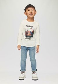 Langärmliges cremefarbenes Shirt mit einem Basketball-Graphic und dem Text "DRIBBLE COURTSIDE CHAMPIONS", kombiniert mit blauen Jeans und weißen Sneakers.