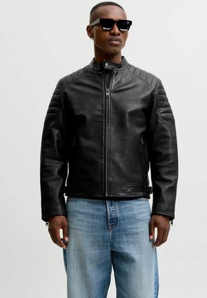 Mees, kes kannab musta nahast biker-jopet, helesiniseid teksapükse, musti päikeseprille ja väikest rõngakujulist kõrvarõngast, ühtlase valge taustaga.