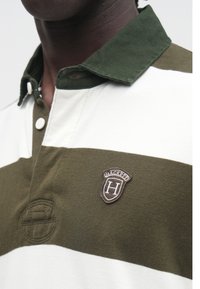 Polo w zielono-białe paski z ciemnozielonym kołnierzykiem. Na materiale znajduje się naszywka z napisem "HACKETT" i okrągłym wzorem.