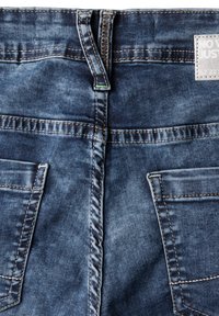 Denim jeans med en mörk tvätt och en urtvättad textur, synlig sömnad och två bakfickor. Tagg högst upp med varumärkesinformation.