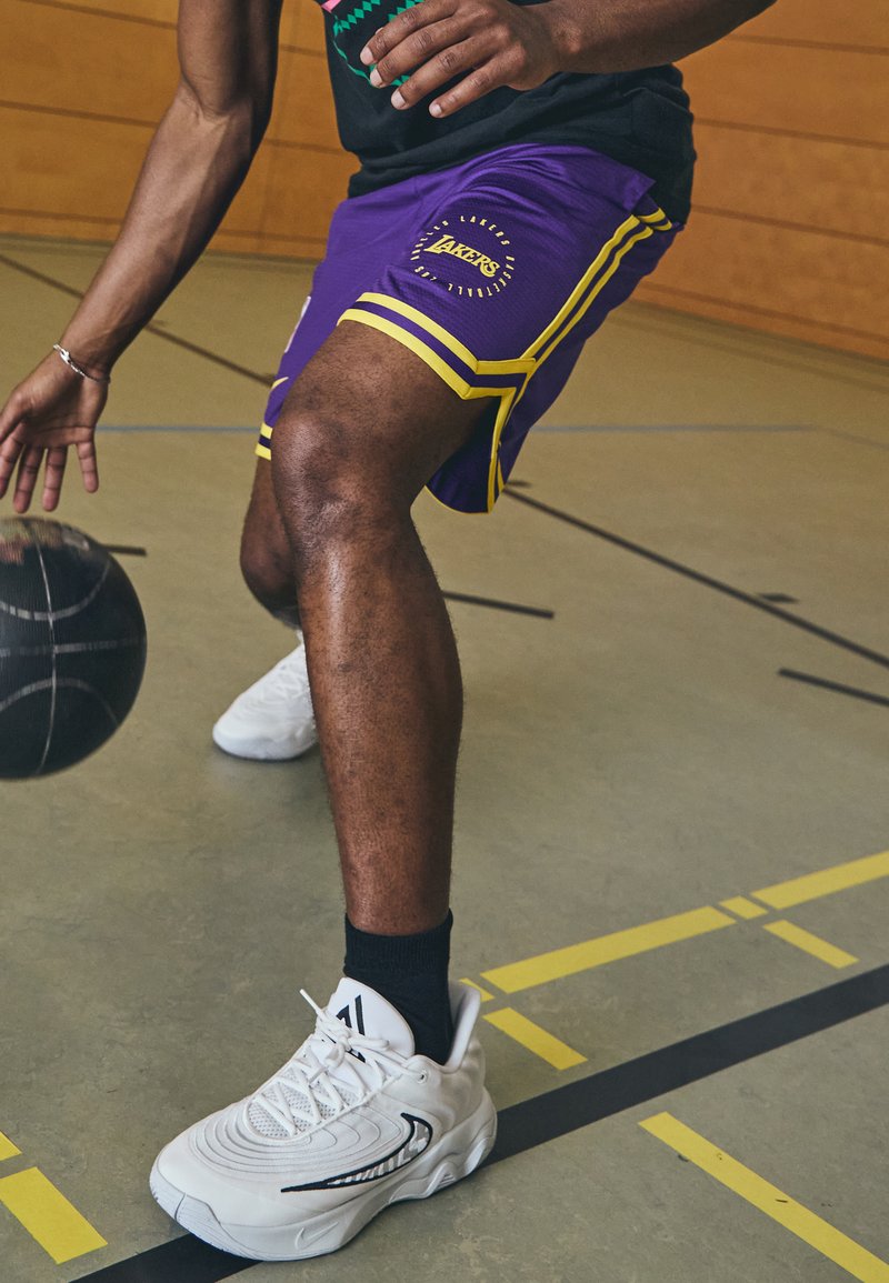 Vijolične košarkarske kratke hlače z rumenimi poudarki in logotipom "Lakers". Bele športne čevlje z črnimi nogavicami, driblajoč črn košarkarski žog na igrišču.