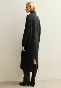 Vrouw met kort blond haar, gekleed in een donkergrijze lange gebreide jurk met een achterkantsplit en zwarte laarzen tot aan de knie, staand opzij.