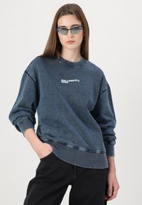 Modra denim majica s rebrastimi manšetami in robom, z logotipom "KARL LAGERFELD JEANS". Nosena z črnimi hlačami, ki prikazujejo sproščen kroj.