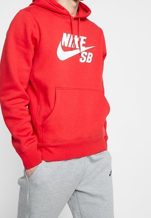Mann trägt roten Nike SB Hoodie mit Fronttasche und hellgraue Nike Jogginghose, Hände in den Taschen, vor einfarbigem Hintergrund.