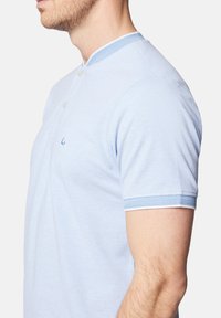 Jasnoniebieska koszulka polo z teksturowanej tkaniny, z kołnierzem henley i dwoma guzikami. Akcenty w nieco ciemniejszym odcieniu niebieskiego na kołnierzu i rękawach.