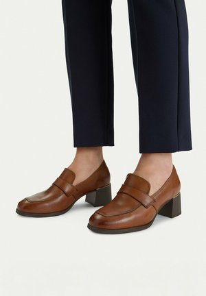 Braune Leder-Loafer mit Absatz, kombiniert mit schwarzer, verkürzter Hose, die die Details der Schuhe und den mittelhohen Blockabsatz hervorhebt.