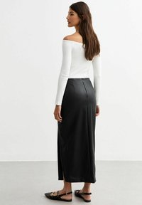 Svart satinmaxikjol med en sidosplit och dragkedjeslutning bak, i kombination med en vit off-shoulder långärmad topp. Platta svarta sandaler.