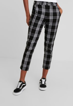 Persoon die zwart-witte geruite cropped broek draagt met handen in de zakken en zwarte sneakers met witte veters op een effen grijze achtergrond.