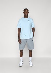 Hele hele sinine Nike t-särk, millel on väike logo ja värviline pritsitud disain, koos hallide lühikeste pükste ja valgete spordijalanõudega.