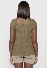 Top verde oliva testurizzato con maniche a sbuffo e dettaglio in vita arricciato. Abbinato a pantaloni corti beige. Il top presenta una vestibilità comoda.