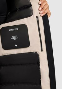 Must ja beež isoleeritud jakki sisemus musta sildiga, millel on kirjas "KHUJOTM" ja "GENUINE BRAND ESTABLISHED 2001", ning tõmblukuga tasku detail.