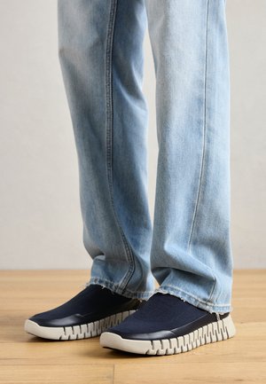 Osoba v světle modrých džínách a tmavě modrých slip-on teniskách s tlustými, texturovanými béžovými podrážkami na dřevěné podlaze.