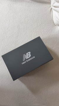 Crna kutija za tenisice New Balance s mat finišem, koja ima srebrni logo na poklopcu i oštre kutne rubove. Kutija leži na teksturiranoj površini.