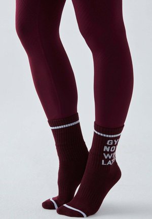 Nogi w bordowych, teksturowanych legginsach i pasujących skarpetkach z białymi paskami i częściowym tekstem na jednolitym tle.