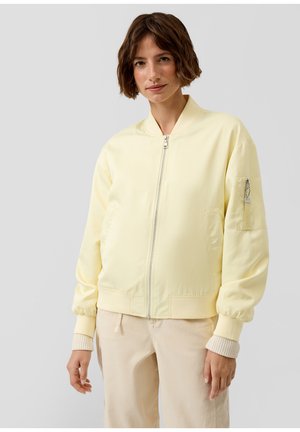 Frau mit kurzen braunen Haaren, die eine hellgelbe Bomberjacke mit Reißverschluss und hellbeige Hose vor einem schlichten Hintergrund trägt.