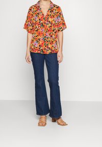 Kurzärmelige, farbenfrohe Bluse mit Blumenmuster in Orange, Pink und Grün, kombiniert mit dunkelbeigen, ausgestellten Jeans und hellbraunen, sandalenähnlichen Absatzschuhen.