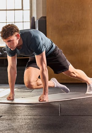 Athletic man in a fitted teal T-shirt and black shorts, demonstrating a workout position on a gray mat in a gym setting.