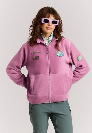 Femme portant une polaire rose zippée avec des écussons et de grandes lunettes de soleil violettes, debout les mains sur les hanches devant un fond uni.