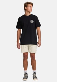 Black cotton t-shirt with a circular graphic on the left chest. Worn with beige shorts and black high-top sneakers. Simple, casual fit.