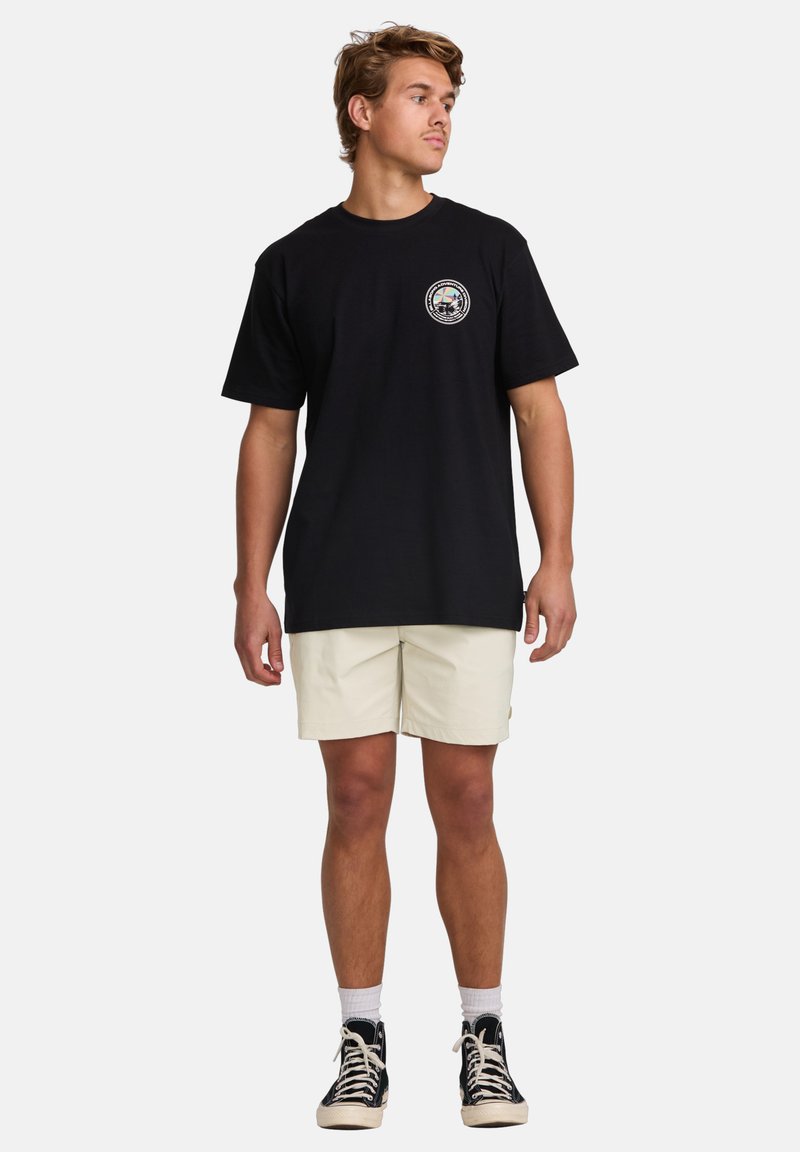 Black cotton t-shirt with a circular graphic on the left chest. Worn with beige shorts and black high-top sneakers. Simple, casual fit.