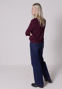 Femme blonde debout, bras croisés, portant un pull bordeaux foncé, un jean bleu à jambes larges et des bottines noires sur un fond uni.