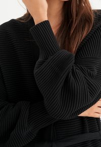 Mustat ribbitetty kardigan, jossa on syvä V-pääntie, ylisuuret hihat ja teksturoitu kangas. Huomionarvoinen pehmeästä, käsittelyystävällisestä suunnittelustaan ja rentosta istuvuudestaan.