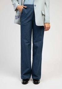 Pantalons en velours côtelé bleu à jambes larges avec des rayures verticales, dotés d'une taille haute et d'un détail à bouton sur le devant, associés à des chaussures en cuir laqué noir.