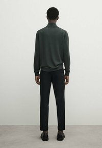 Pull vert foncé à coupe décontractée, manches longues et col montant, associé à un pantalon noir et des chaussures noires, vu de dos.