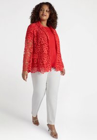 Giacca rossa in pizzo con motivo floreale, design a strati, indossata sopra una maglietta rossa e pantaloni bianchi. Tacchi argentati completano l'outfit.
